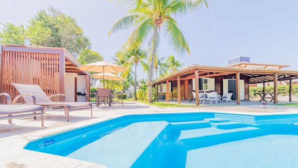 A swimming pool with lounge chairs and umbrellas, surrounded by wooden structures and palm trees. - GB69 Excelente Casa a 400m da Praia (Camaçari)