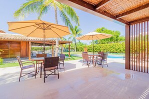 An outdoor patio with wicker furniture and large umbrellas, a pool in the background, and a wooden fence. - GB69 Excelente Casa a 400m da Praia (Camaçari)