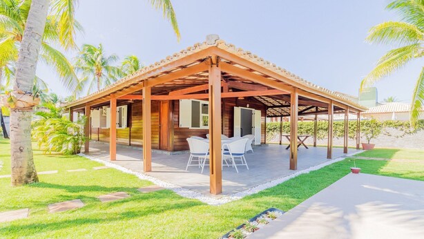 A covered outdoor area with a wooden structure, white chairs, and a small table. - GB69 Excelente Casa a 400m da Praia (Camaçari)