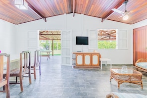 A spacious room with a high ceiling, a television mounted on the wall, a dining area with wooden chairs, a wicker sofa, and a stone-tiled floor. - GB69 Excelente Casa a 400m da Praia (Camaçari)