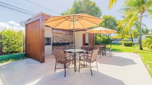 An outdoor patio area with a round table and chairs, two large umbrellas, and a grill station. - GB69 Excelente Casa a 400m da Praia (Camaçari)