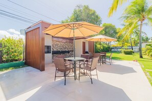 An outdoor patio area with a round table and chairs, two large umbrellas, and a grill station. - GB69 Excelente Casa a 400m da Praia (Camaçari)