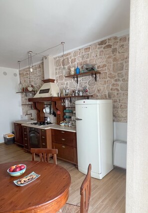 A compact kitchen with a stone wall, a white refrigerator, wooden cabinets, and a dining table with a bowl of fruit.