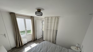 A bedroom with a bed, a window with curtains, a door, and a ceiling fan. - Petite Maison au Calme Dans un Village Typique (Saint-Pierre-d'Oléron)