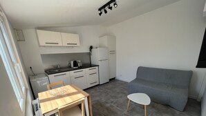 A compact living space with a kitchenette, a dining table with chairs, a gray sofa, and a small white table. - Petite Maison au Calme Dans un Village Typique (Saint-Pierre-d'Oléron)
