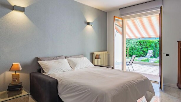 Une chambre à coucher avec un lit, une lampe de chevet et une vue sur un jardin, visible à travers une porte ouverte.