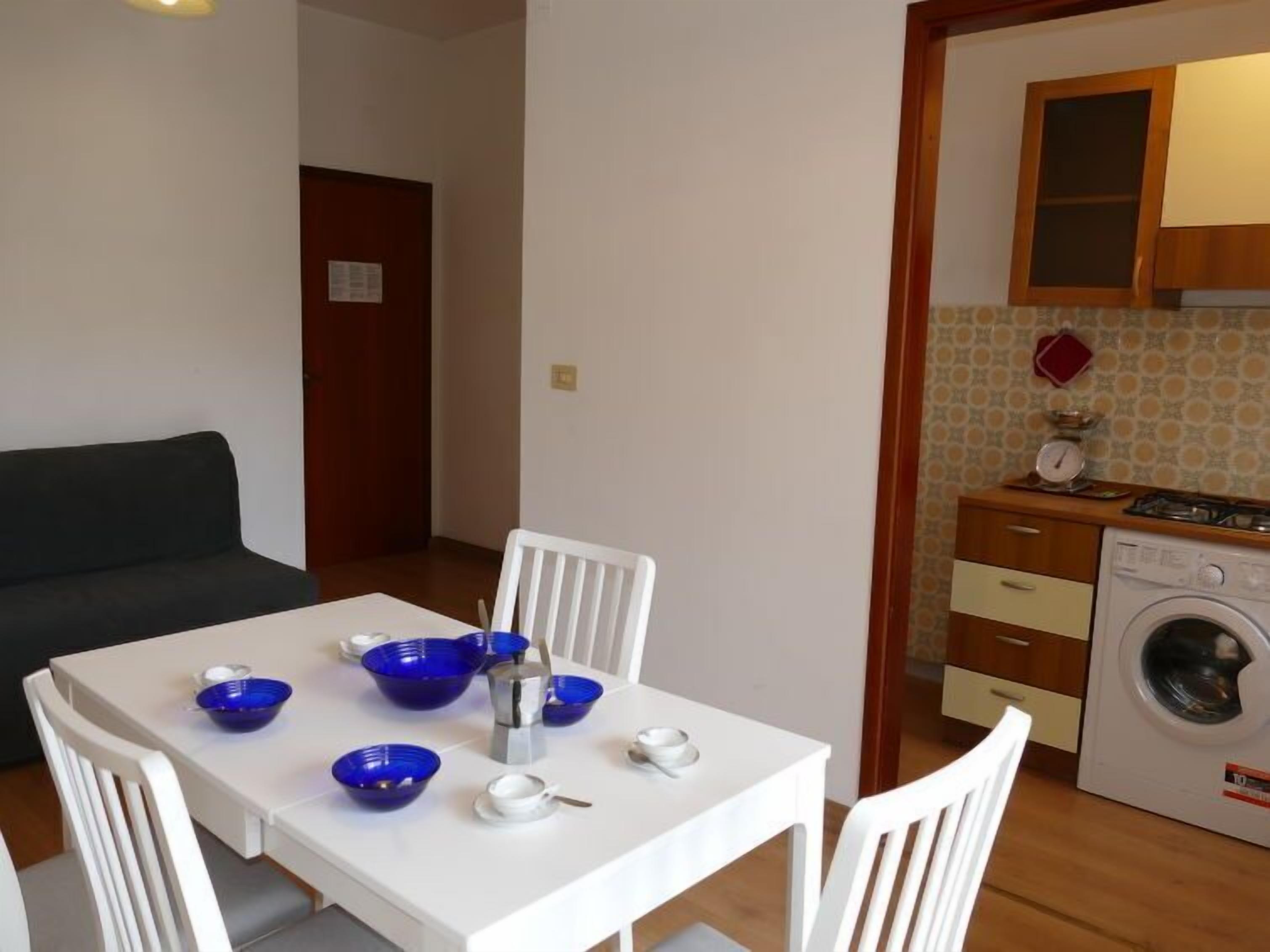 A compact living space with a dining table set for four, a sofa, a washing machine, and a kitchen area with a stove and cabinets.