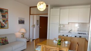 Ein modernes Wohnzimmer mit einer Couch, einem Esstisch mit Stühlen, einer Küchenzeile mit weißen Schränken und einem Flur, der in einen weiteren Raum führt.