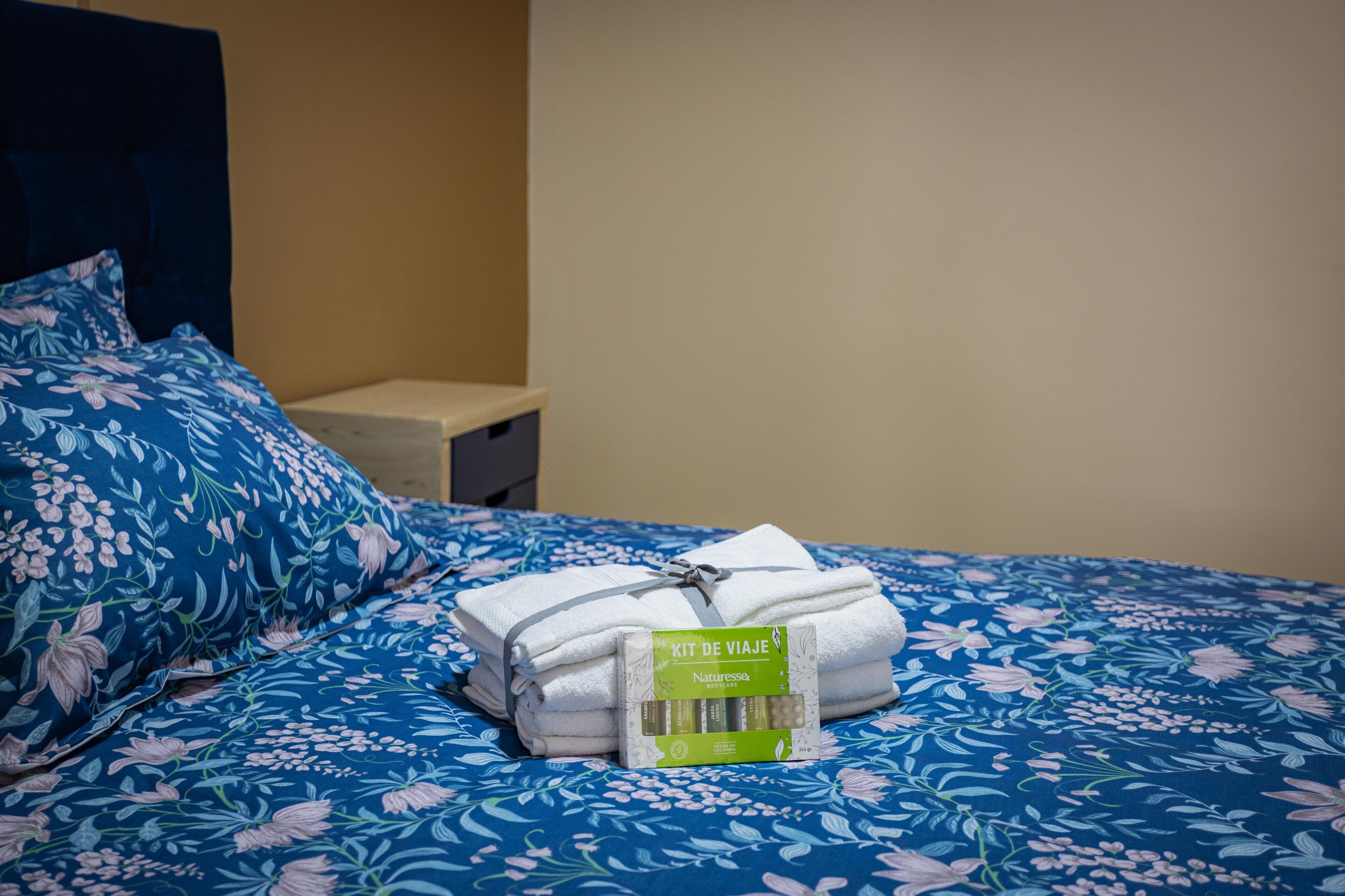 A neatly made bed with a floral bedspread, a set of white towels, and a green package labeled "KIT DE VIAJE" on a hotel room bed.