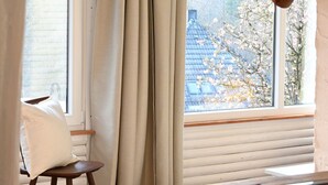 Ett rum med utsikt över ett hus och blommande träd genom ett fönster med träpersienner.