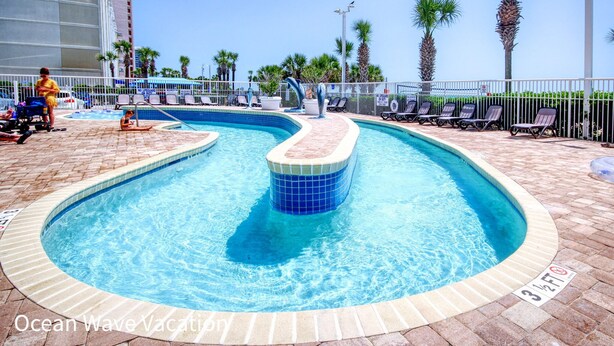 A swimming pool with a curved design, surrounded by lounge chairs and palm trees. - Amazing Beach Condo with Water Park at Dunes Village Resort (Myrtle Beach)