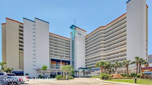 A multi-story hotel with a central tower, multiple balconies, and a parking area with palm trees. - Amazing Beach Condo with Water Park at Dunes Village Resort (Myrtle Beach)