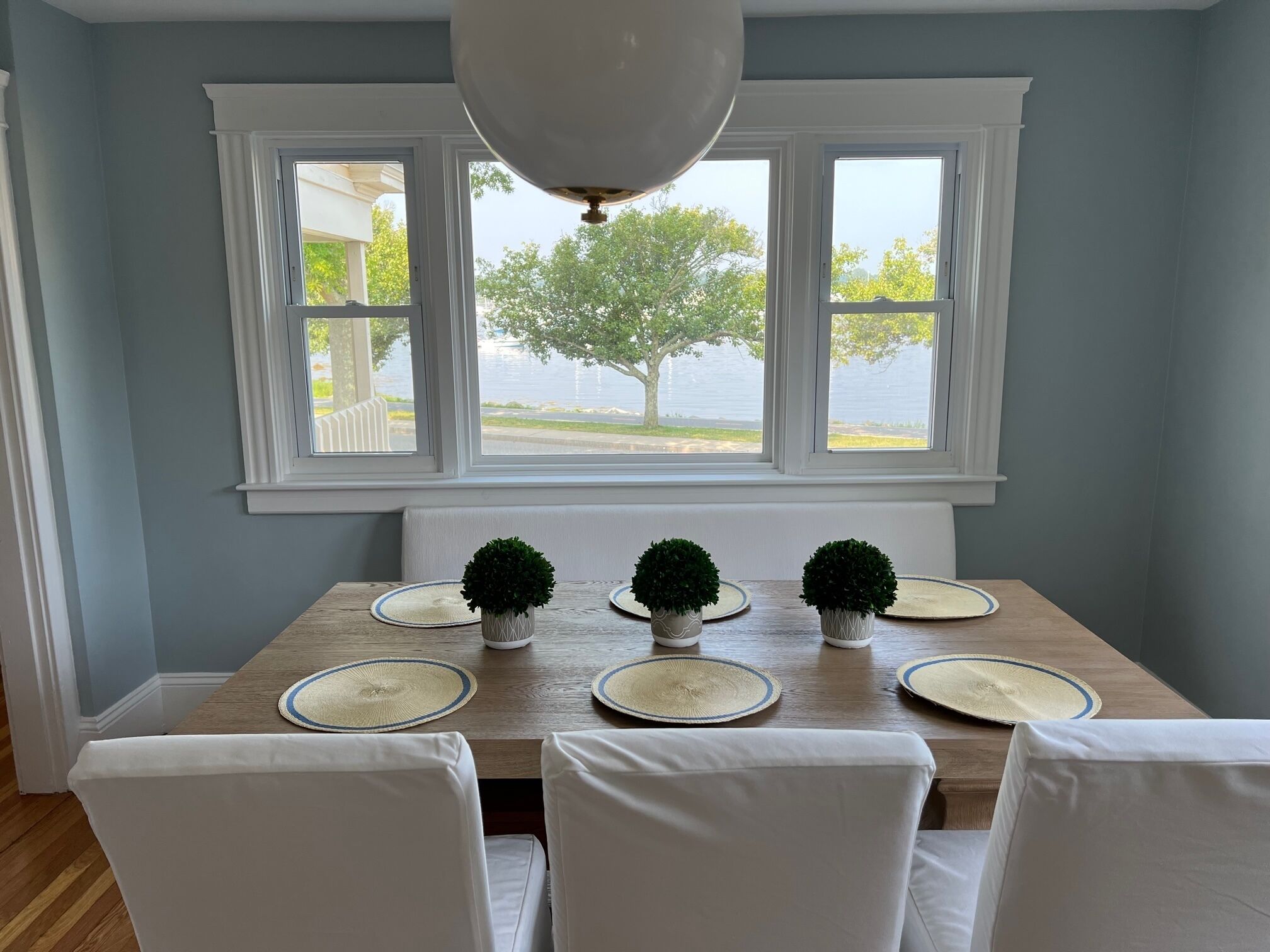 Un coin repas avec une table en bois, des chaises blanches et une vue sur un lac, visible à travers de grandes fenêtres.
