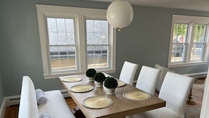 Un coin repas avec une table en bois, des chaises blanches et un banc. Il y a deux fenêtres avec des cadres blancs et un luminaire blanc au plafond.
