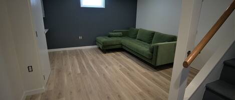 A room with a green sectional sofa, wooden flooring, a small window, and a staircase.