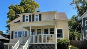 Une maison à deux étages avec un porche à l’avant, un escalier menant au deuxième étage et deux chaises Adirondack dans le jardin.