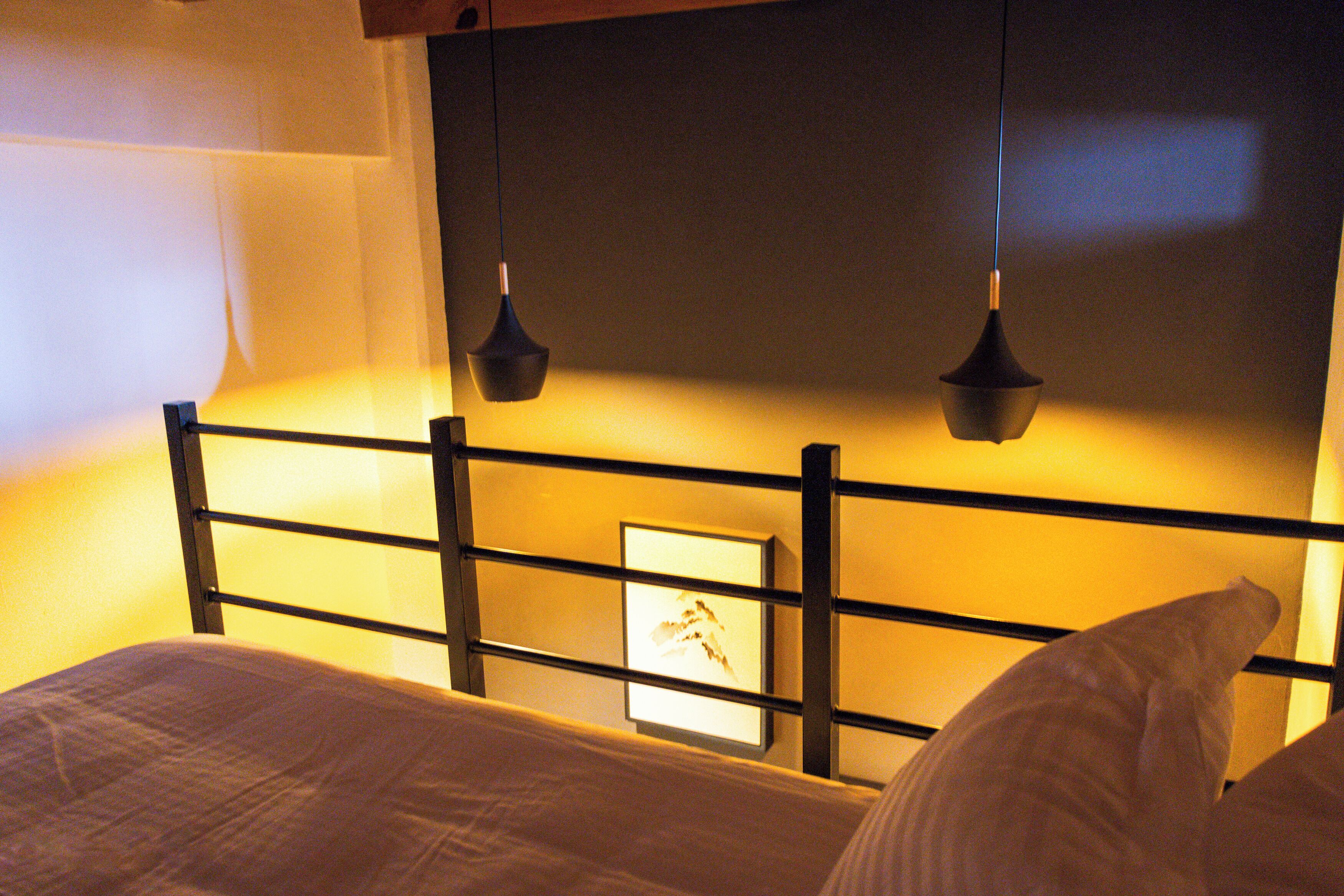 A neatly made bed with a headboard featuring a framed picture, a bedside table, and two hanging pendant lights.