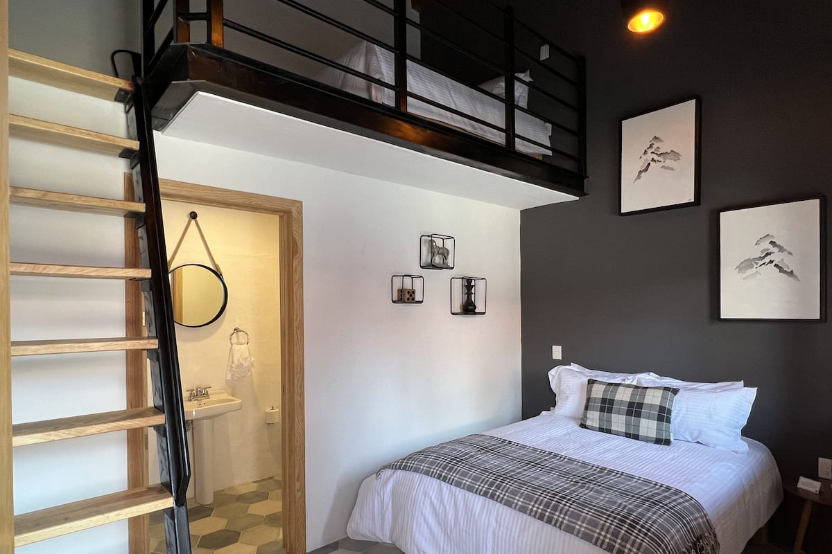 Um quarto com cama, uma escada de madeira, um banheiro pequeno e uma escada que leva a um andar superior.