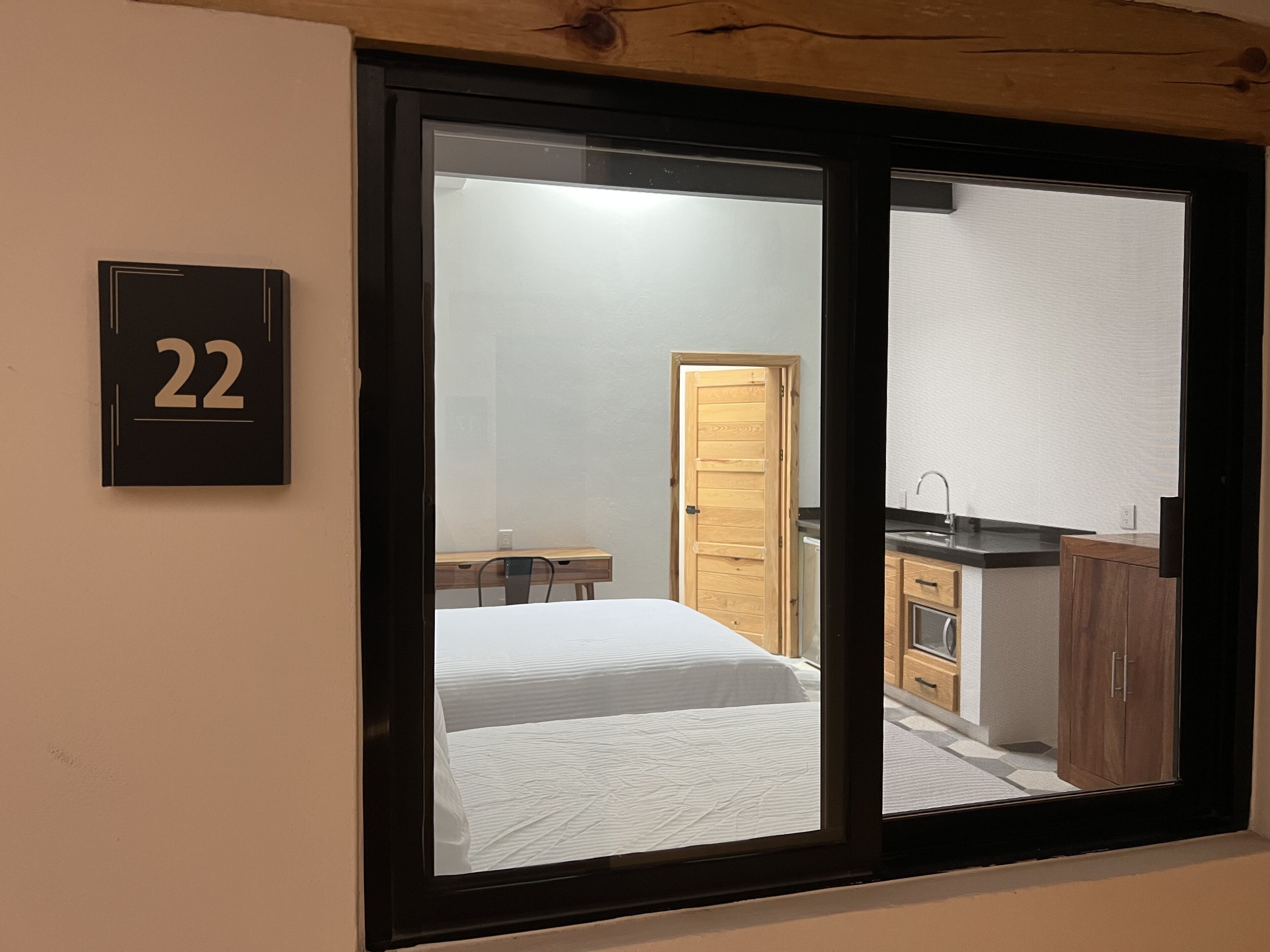 A modern room with a large sliding glass door leading to a kitchen area. A wooden door is visible inside the room, and a small wooden table with chairs is placed near the door. The room has a number '22' sign on the wall.