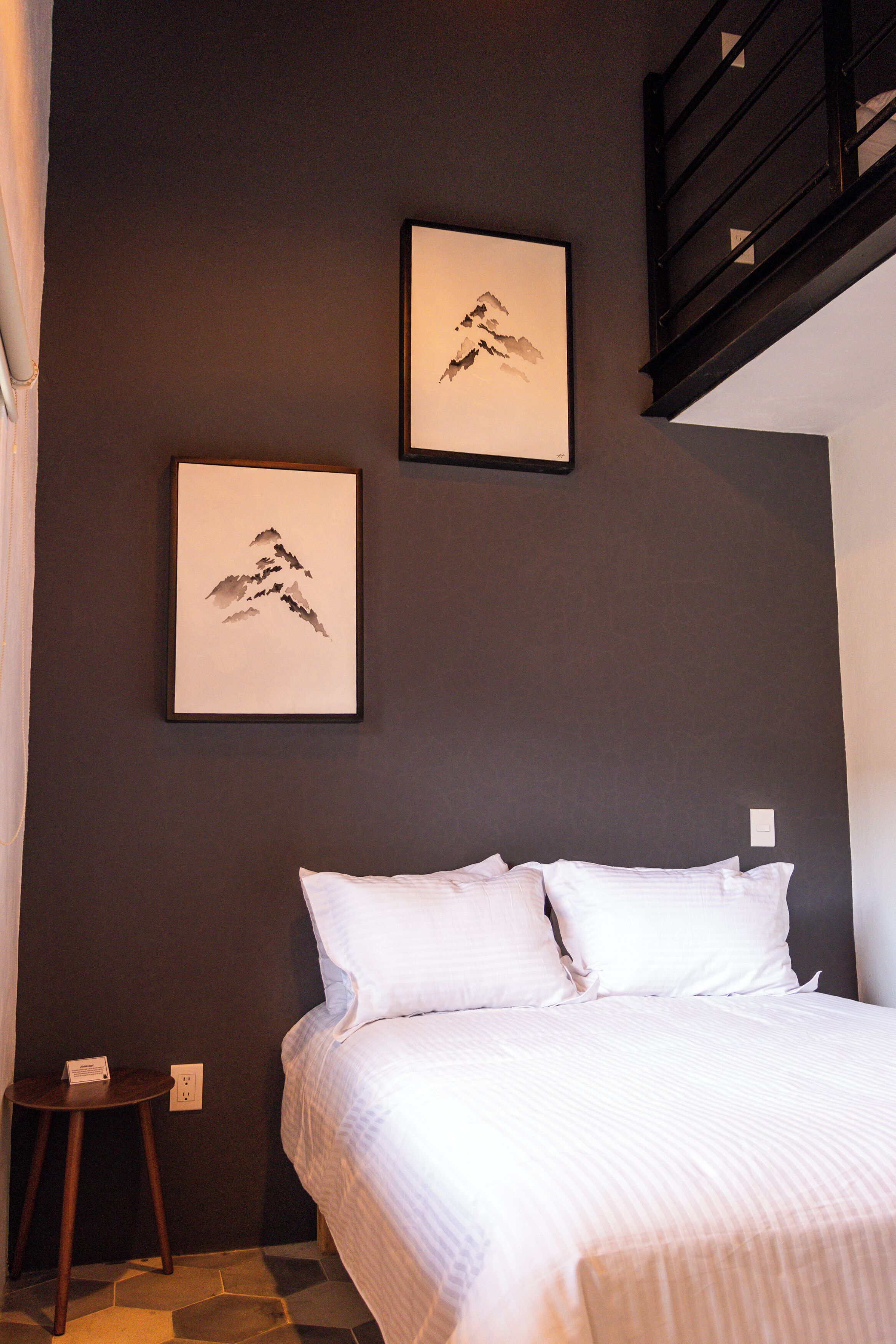 Un dormitorio con una cama, dos cuadros en la pared, un taburete de madera y una escalera.