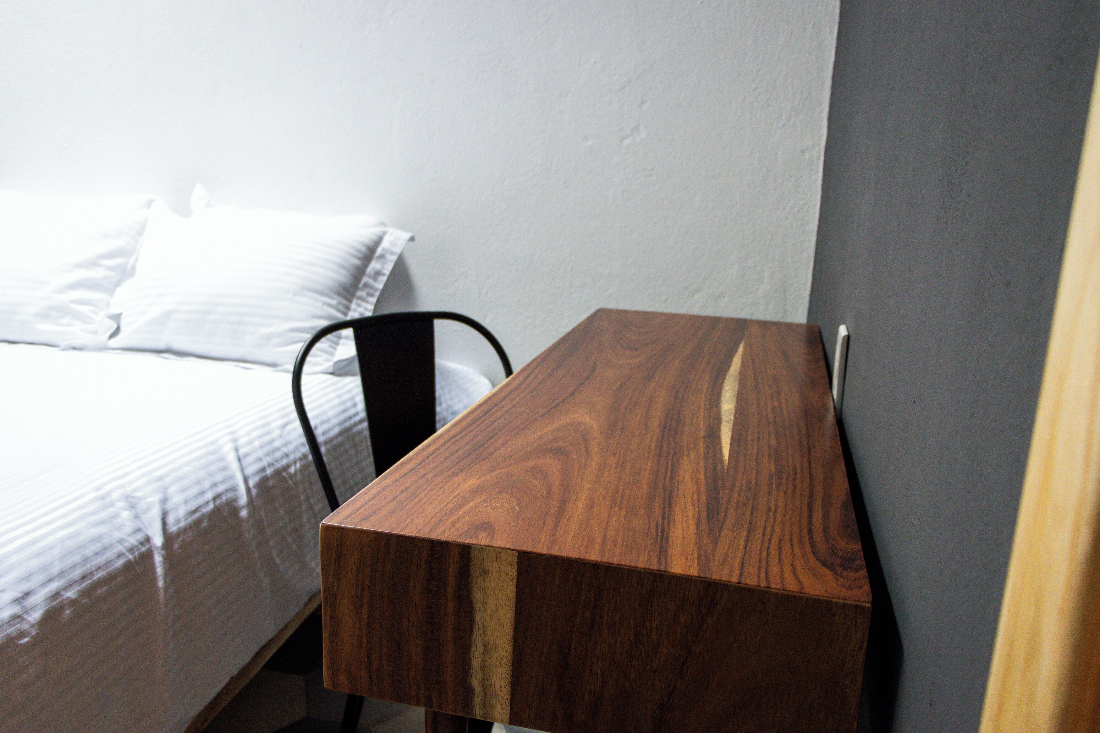 A wooden desk with a chair, a bed with white bedding, and a gray wall.