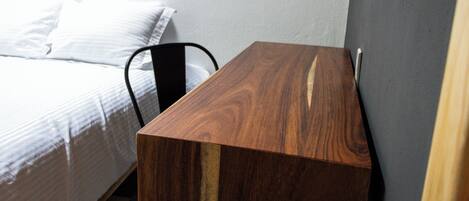 Un escritorio de madera con una silla, una cama con ropa blanca y una pared gris.