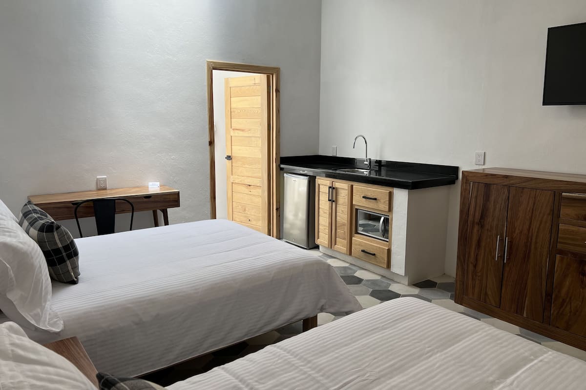 Quarto compacto com duas camas, uma mesa pequena, uma porta de madeira, cozinha compacta e uma TV fixada na parede.