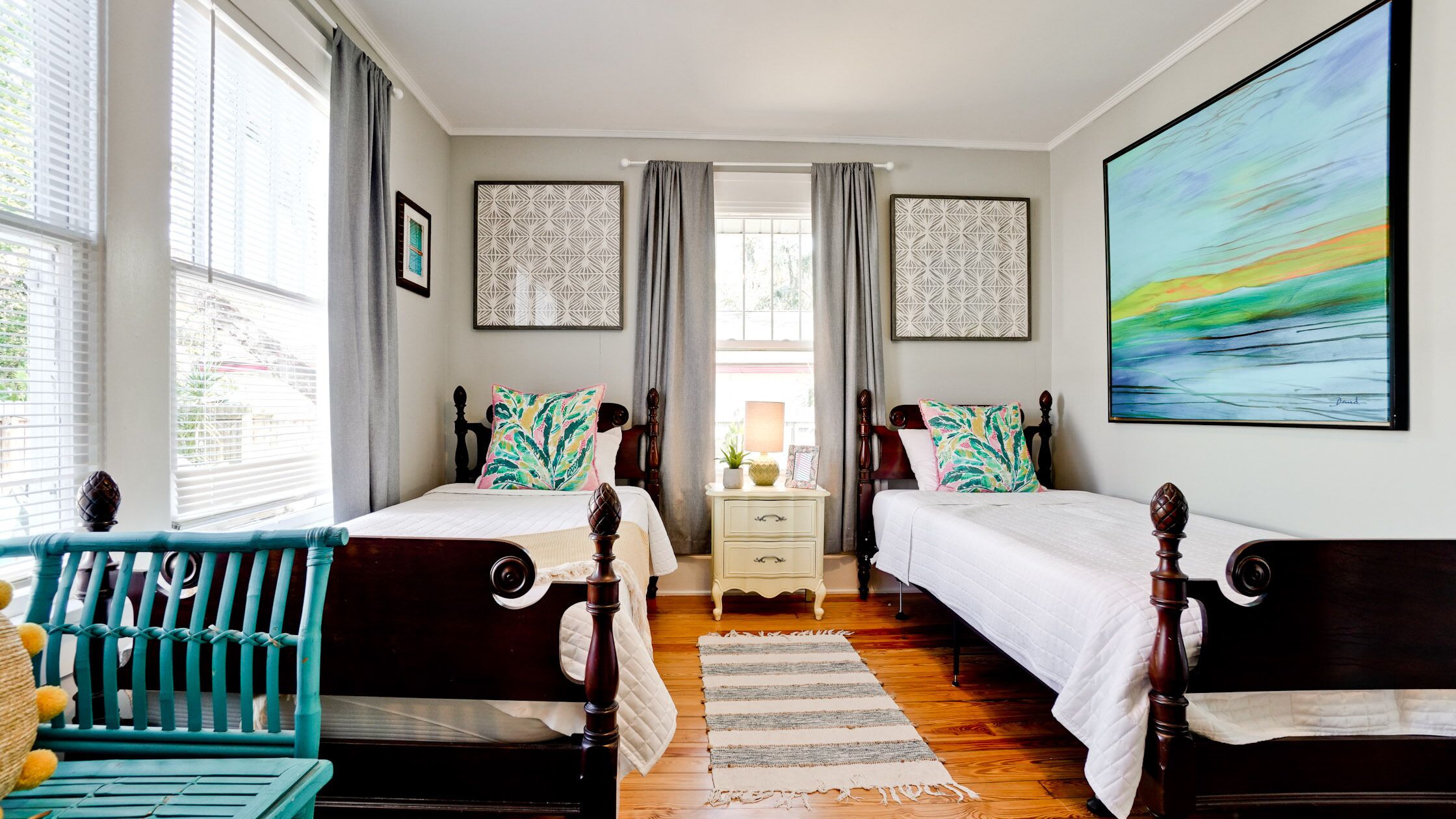Une chambre avec deux lits, une table de chevet, une commode, une fenêtre avec des rideaux et un grand tableau au mur.