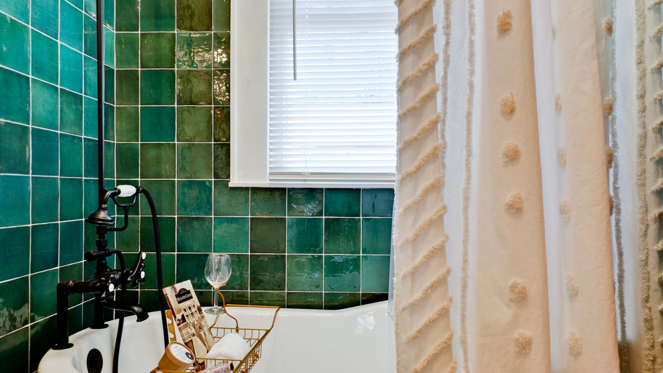 Un bagno con piastrelle verdi, una vasca da bagno, una finestra con persiane e un portasapone con articoli da toilette.