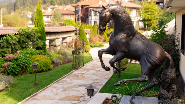 A bronze horse statue in a garden with a stone pathway, surrounded by greenery and houses in the background. - Armaco Residence (Koprivshtitsa)