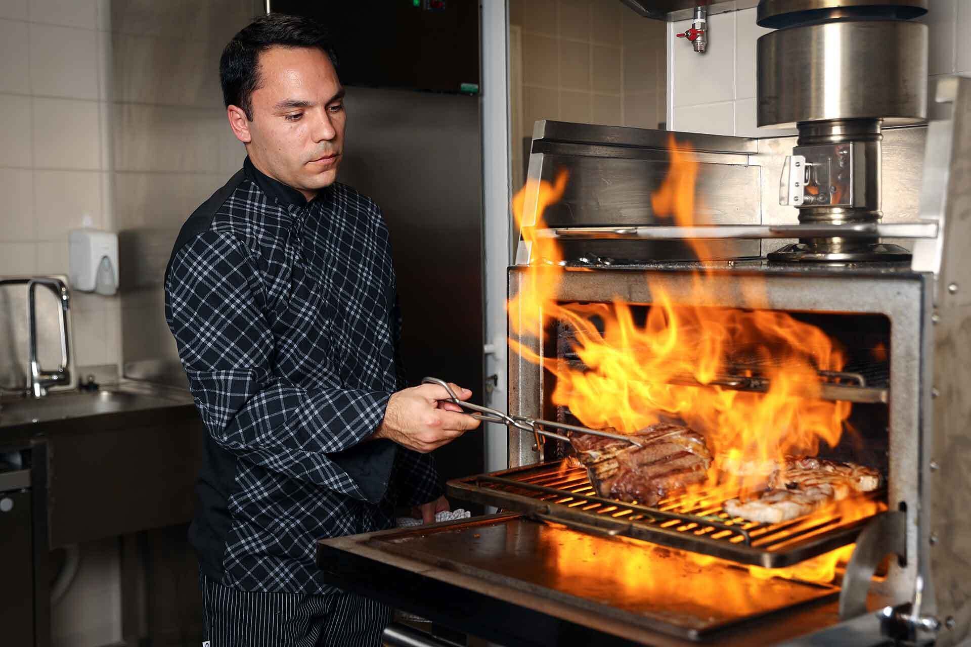 Un chef asando carne en una parrilla de alta temperatura en una cocina.