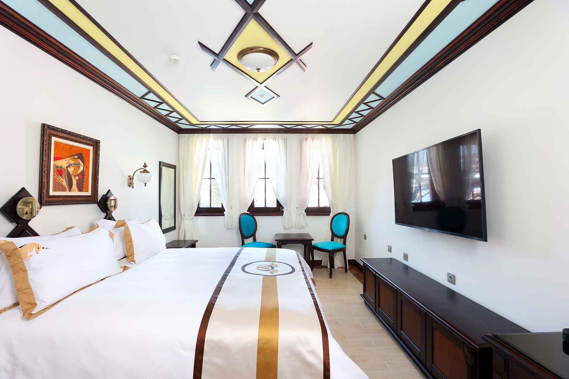 A hotel room with a large bed, a TV, two chairs, a small table, and a painting on the wall.