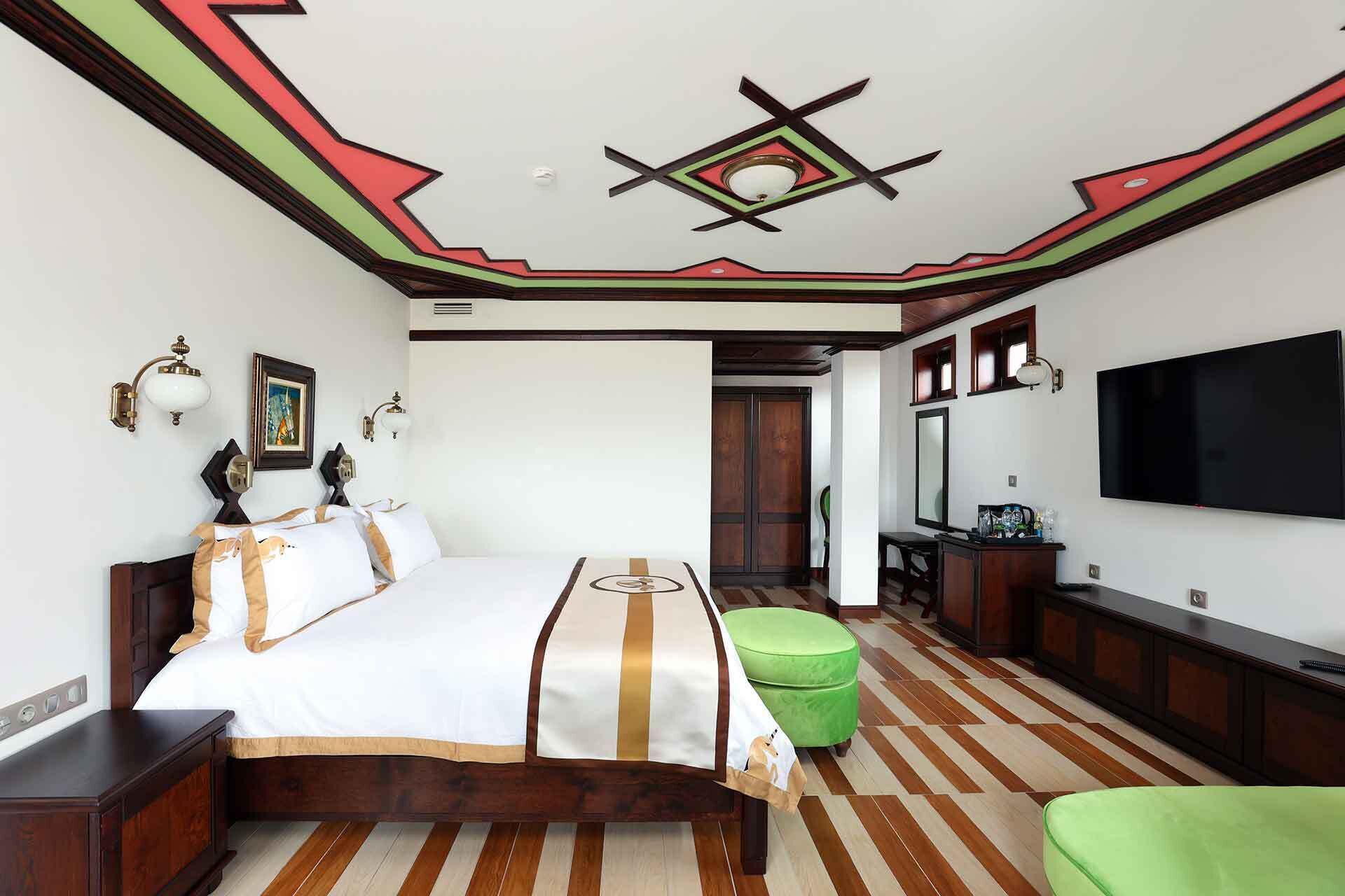 A hotel room with a bed, a green ottoman, a TV, and a wooden floor.