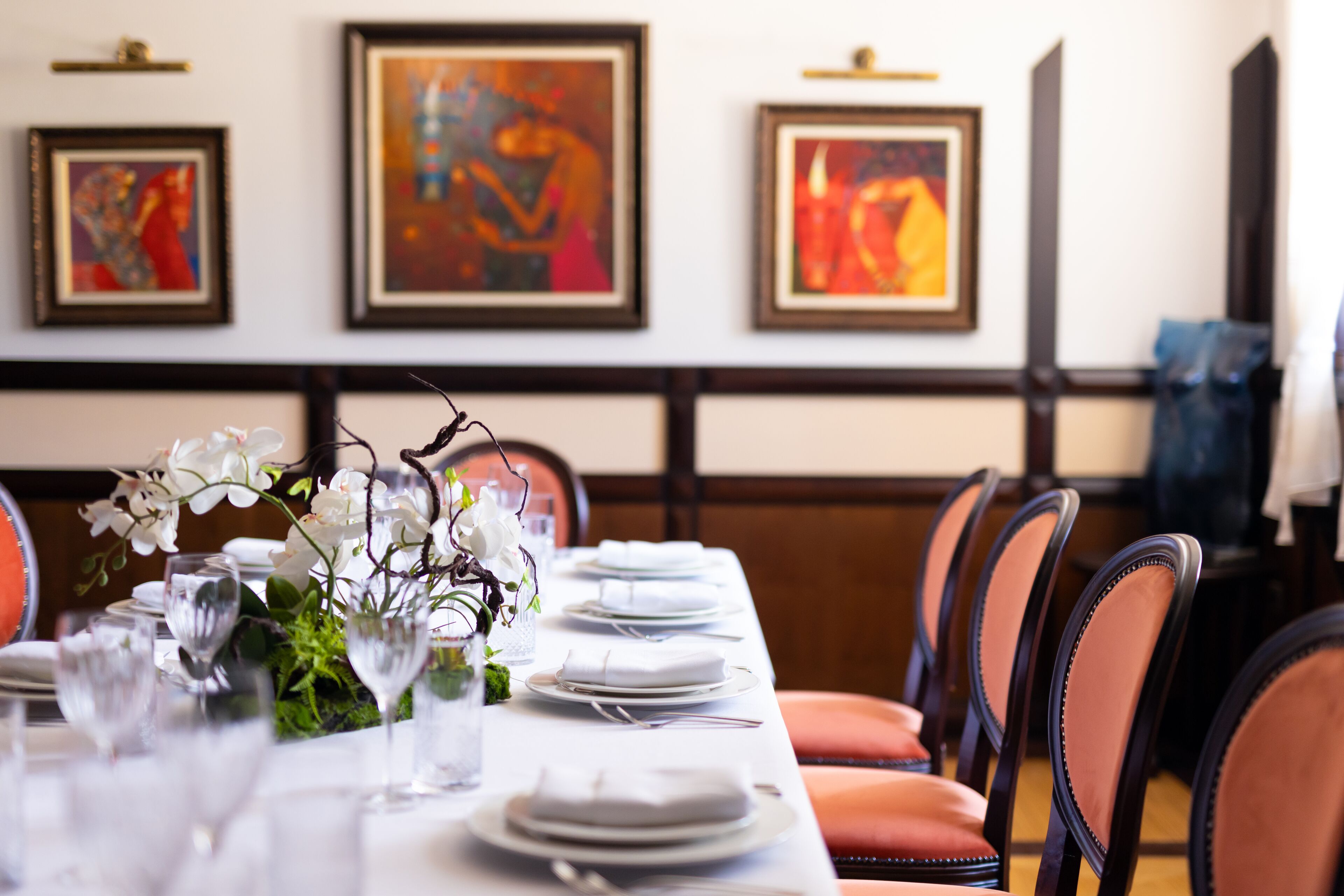 A dining table set with white plates, glasses, and a floral centerpiece. There are framed artworks on the wall behind the table.