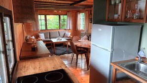 A wooden cabin interior with a dining area, kitchen, and a large window overlooking greenery. - Charmante Cabane en Pleine Nature (Rhodes)