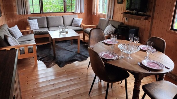 A cozy cabin interior with a wooden dining table set for four, a sofa with cushions, and a television mounted on the wall. - Charmante Cabane en Pleine Nature (Rhodes)