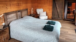 A wooden cabin bedroom with a bed, a chair, a nightstand, and a hanging light fixture. - Charmante Cabane en Pleine Nature (Rhodes)