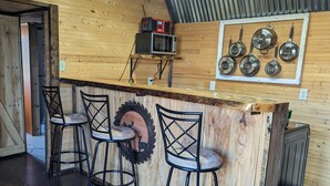 Un área de bar rústica de madera con cuatro taburetes, una rueda de neumático montada y una pared que exhibe varias ollas y sartenes.