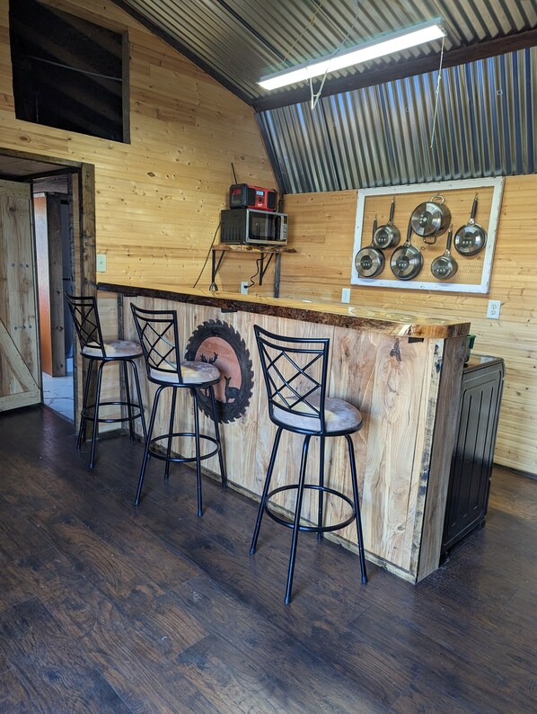 Un área de bar rústica de madera con cuatro taburetes, una rueda de neumático montada y una pared que exhibe varias ollas y sartenes.