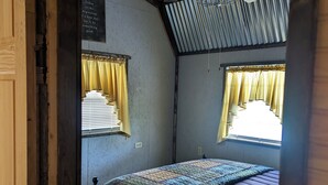 Una habitación pequeña y rústica con una cama, un ventilador de techo y dos ventanas con cortinas.