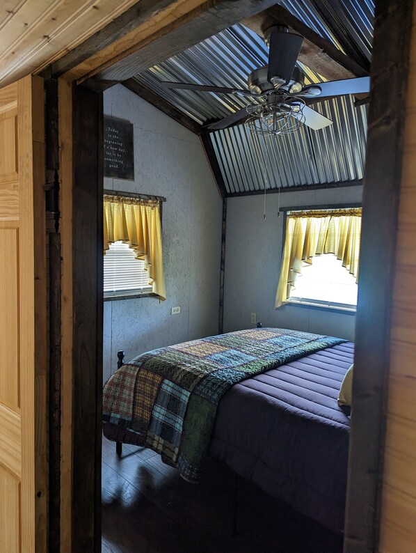 Una habitación pequeña y rústica con una cama, un ventilador de techo y dos ventanas con cortinas.
