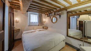 Een slaapkamer met een groot bed, houten balken in het plafond, een nachtkastje, een spiegel en een raam met gordijnen.