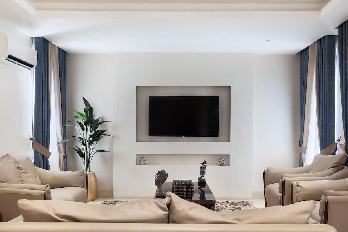 A modern living room with a flat-screen TV mounted on the wall, beige sofas, a potted plant, and a fireplace with decorative items.