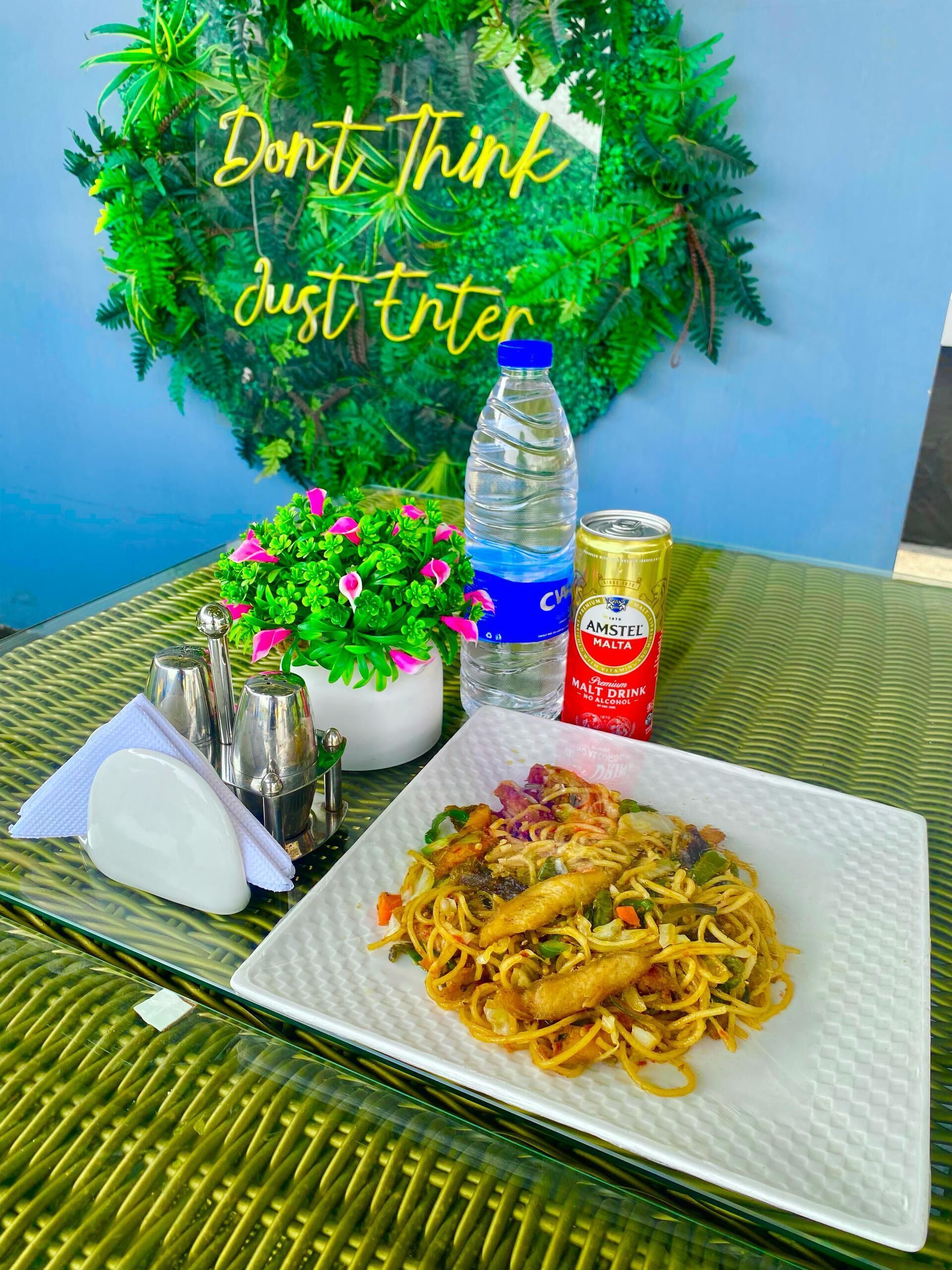 A table with a plate of noodles, a bottle of water, a can of Amstel beer, a small plant, and condiments.
