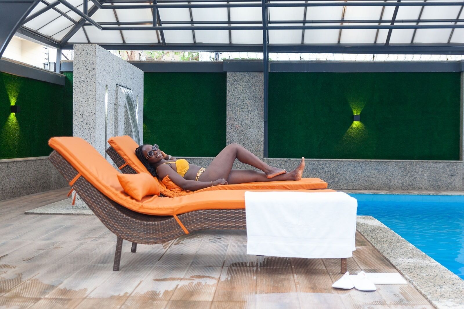 A person relaxing on a sun lounger by a pool with a glass roof and green walls.