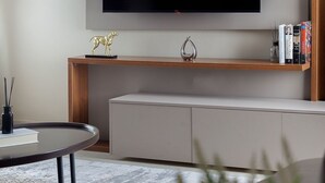 Un soggiorno moderno con una grande TV a schermo piatto montata a parete, una mensola in legno con oggetti decorativi, un divano grigio e un tavolino rotondo con un libro e una candela.