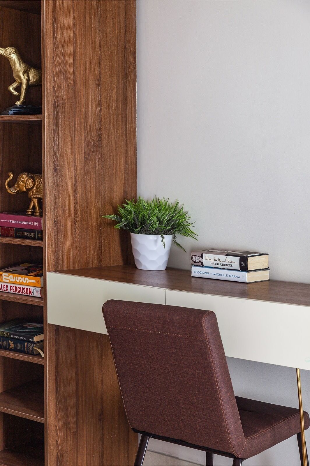 Une bibliothèque en bois agrémentée d’objets décoratifs, un bureau blanc avec des livres et une chaise marron.