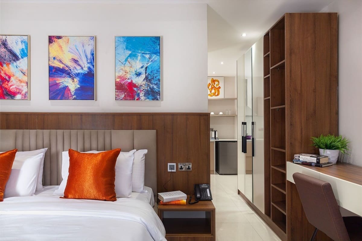 A modern hotel room with a large bed, a desk, and a wardrobe. There are abstract paintings on the wall and a small plant on the desk.