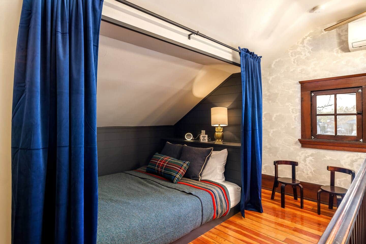 Ein Schlafzimmer mit geneigter Decke, einem Bett mit blauer Decke, einem Nachttisch mit Lampe und einem Fenster mit Blick.