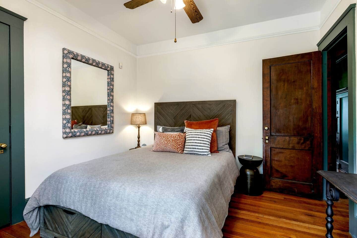 A bedroom with a bed, a nightstand, a lamp, a mirror, and a wooden door.